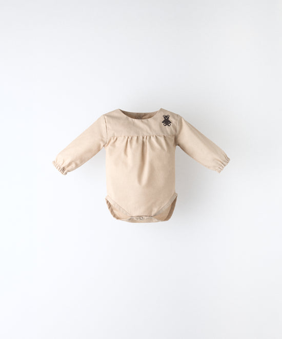 Delantero del body Liberty en manga larga y color arena blanqueada. Confeccionado en 100 % algodón orgánico certificado GOTS y oso de Kala bordado en canesú. Suavidad y cuidado de la piel para tu bebé. www.kala.es