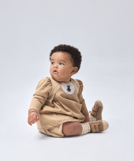 Modelo bebé vestido con El Conjunto Harper en color avellana en combinación de sarga marrón 100 % algodón GOTS y pana beige de algodón BCI; forro 100 % algodón. y cubrepañal en pana beige de algodón BCI (97 %) + 3 % elastano; forro 100 % algodón. Elegancia y diseño para tu bebé. www.kala.es