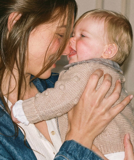 Bebé en brazos con chaqueta de punto beige de bodoques y cuello Príncipe de Gales
