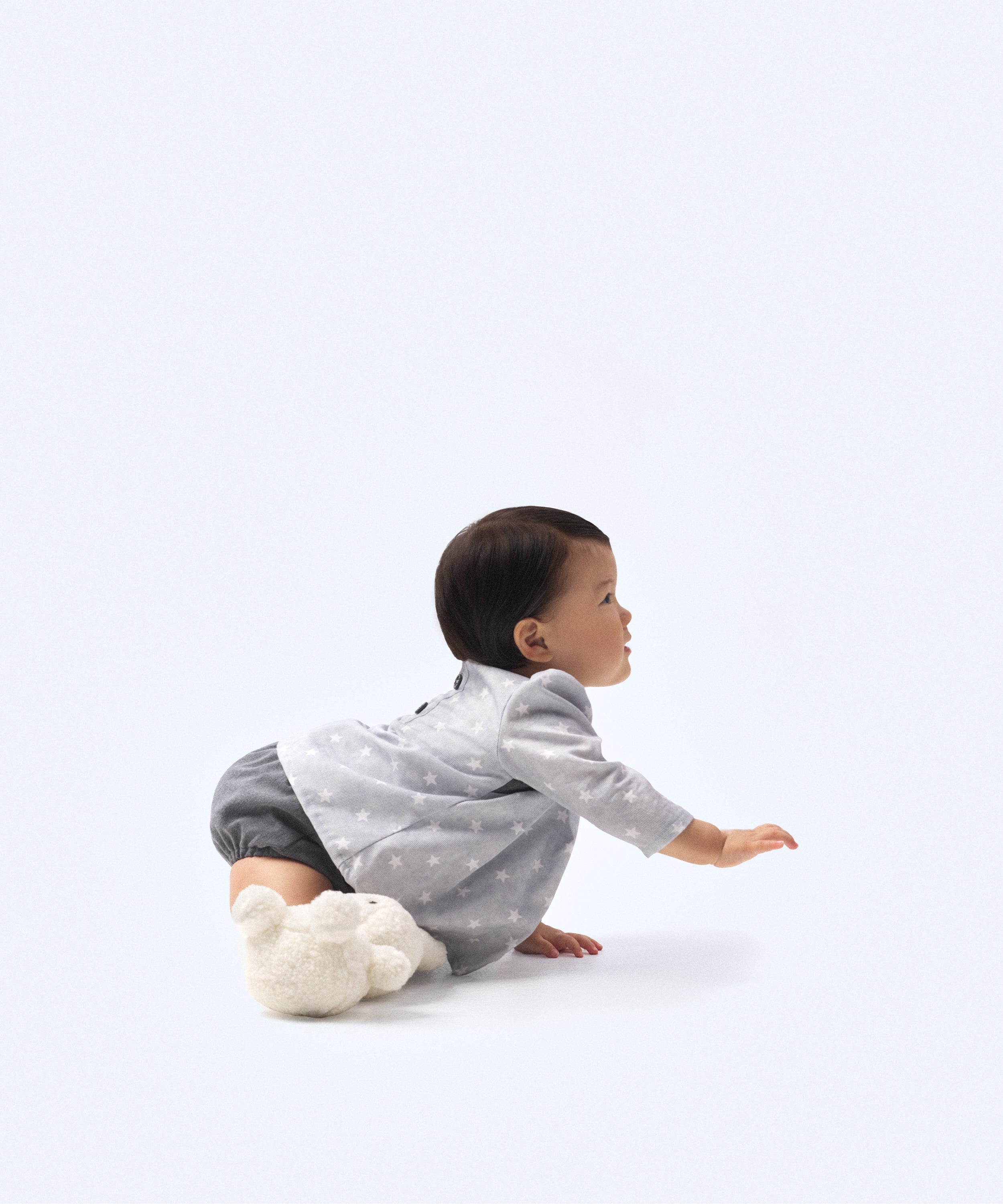 Look de ropa infantil de bebé de Kala. La bebé lleva puesto El conjunto Harper de tejido estampado de estrellas blancas con fondo en color gris bebé y sarga denim azul grisáceo. Compuesto por blusón con manga larga y cubrepañal. Sofisticación y elegancia para la infancia. 