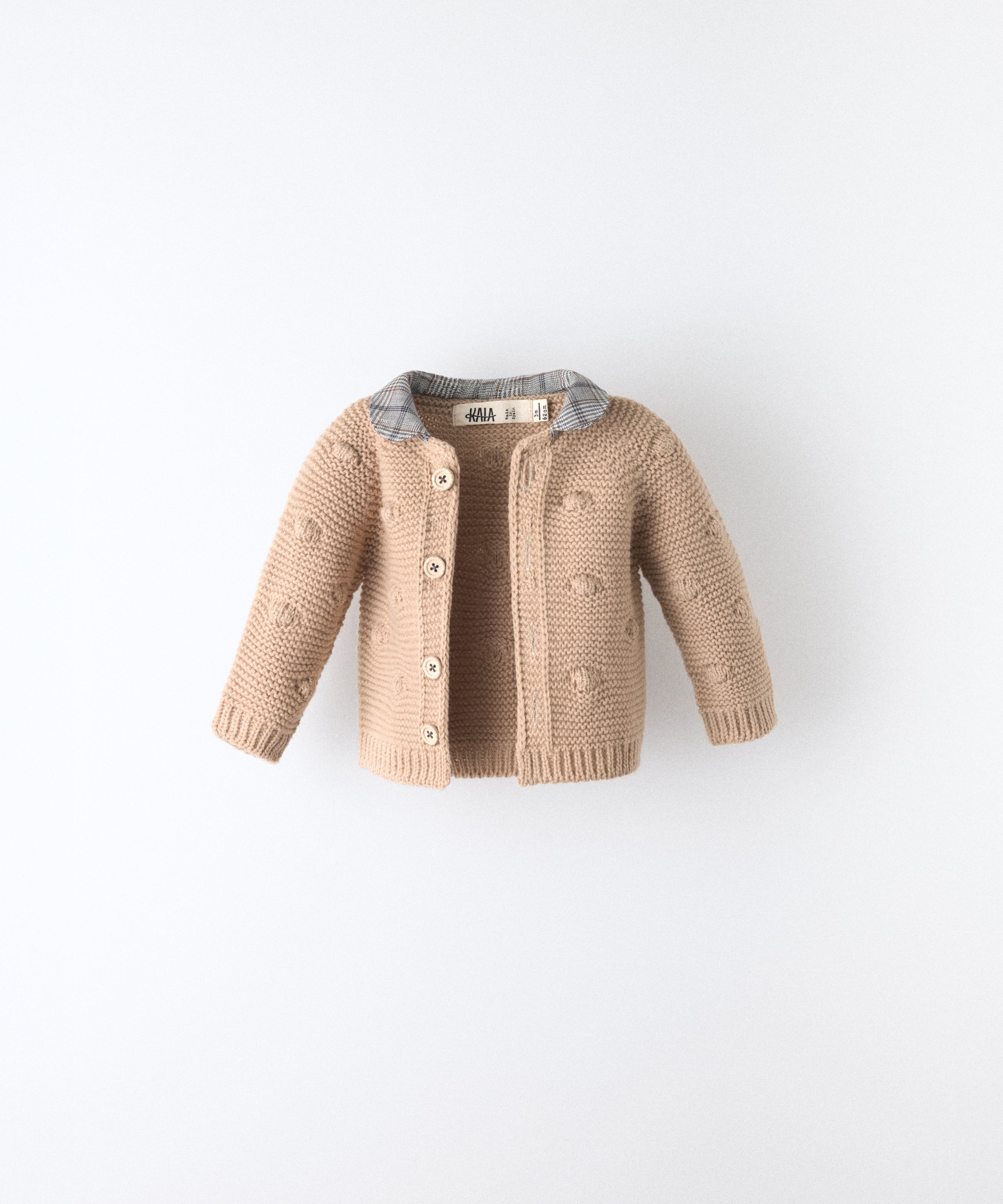 La Chaqueta Brunel beige en punto acrilico con bodoques, botones OEKO TEX y cuello contrastado con tejido cuadros con certificación GRS. Ideal para abrigar a tu bebé. www.kala.es