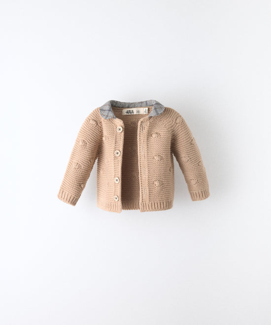 La Chaqueta Brunel beige en punto acrilico con bodoques, botones OEKO TEX y cuello contrastado con tejido cuadros con certificación GRS. Ideal para abrigar a tu bebé. www.kala.es