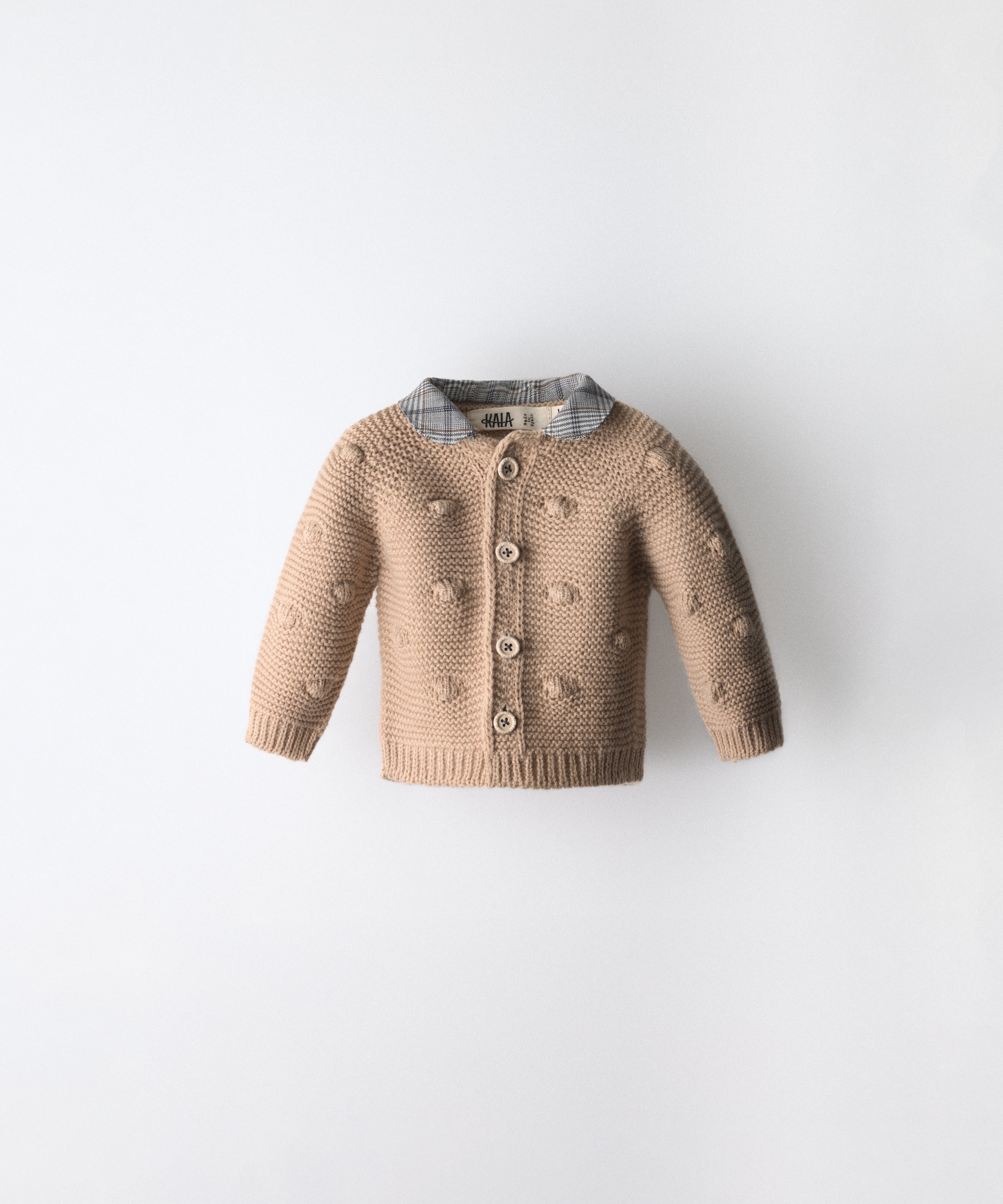 La Chaqueta Brunel beige en punto acrilico con bodoques, botones OEKO TEX y cuello contrastado con tejido cuadros con certificación GRS. www.kala.es