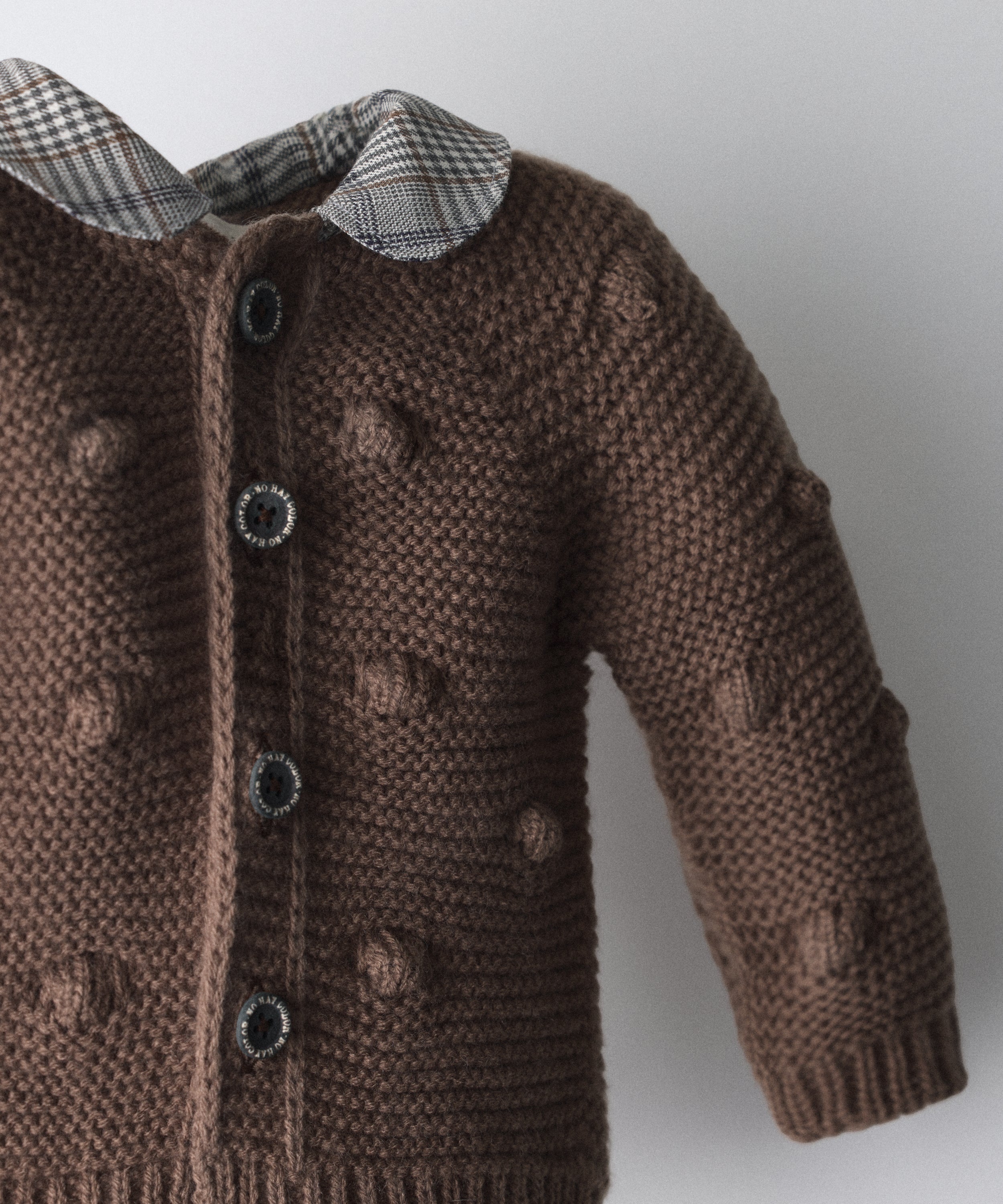 Detalle textura de La Chaqueta Brunel marrón en punto acrilico con bodoques. Cuello contrastado con tejido cuadros con certificación GRS. Esta chaqueta de bebé celebra la unión entre tradición y diseño contemporáneo.www.kala.es