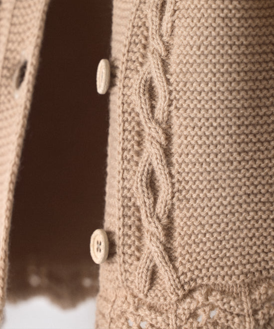 Detalle de trenzas en relieve en el delantero al tono de La Chaqueta Jolly en punto acrílico y color beige. Cierre con botones OEKO TEX al tono. Elegancia y artesanía para tu bebé. 