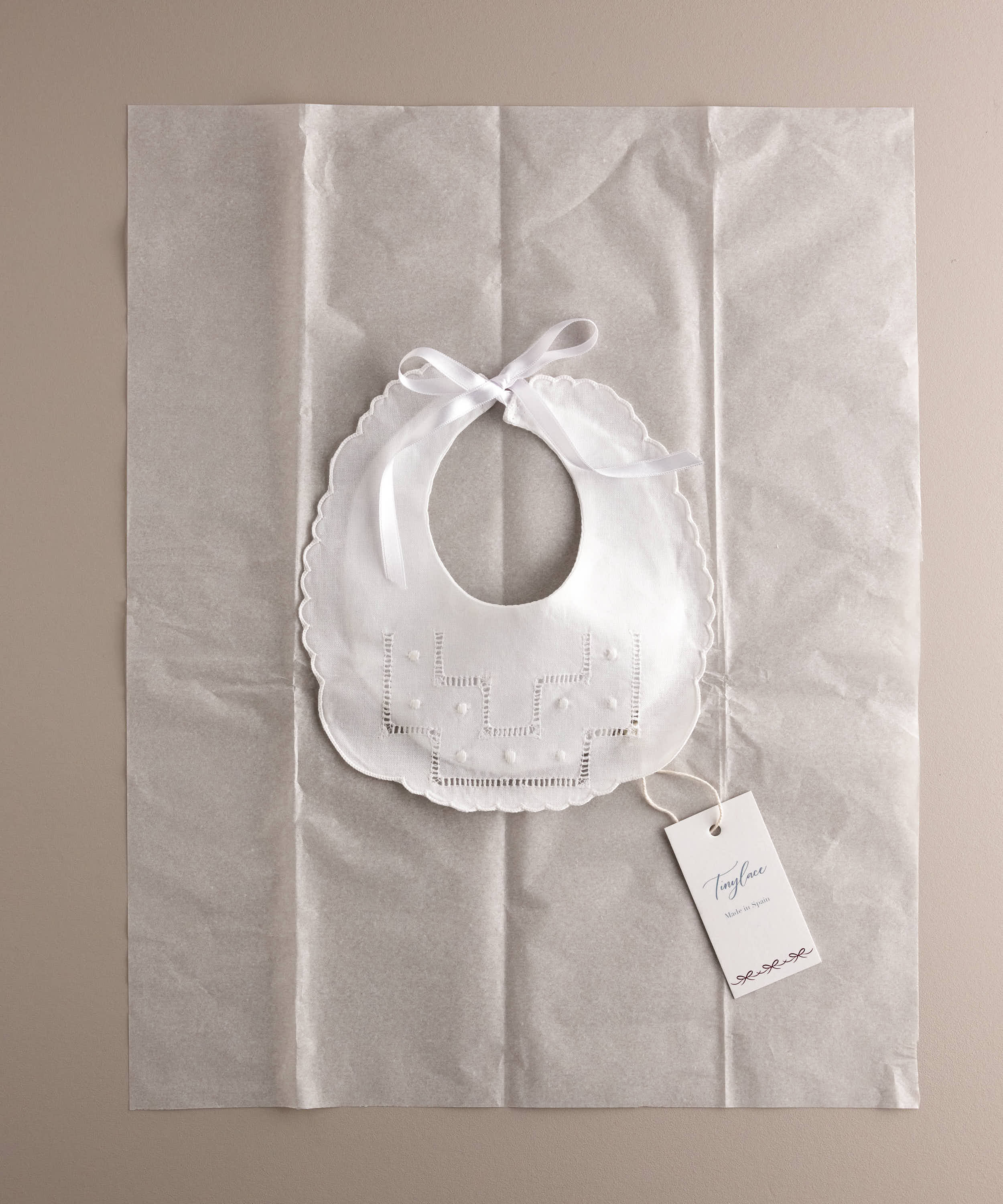 Babero recién nacido de algodón bordado con escalera de vainica y bodoque sobre papel seda y etiqueta marca a modo de packaging. Ref 8648W