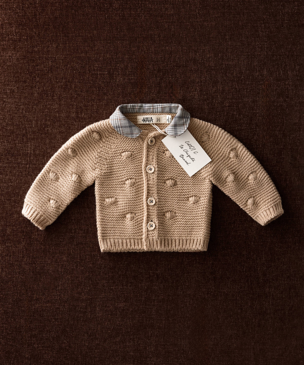 Chaqueta de punto de bebé beige con bodoques de punto y cuello Príncipe de Gales sobre fondo marrón