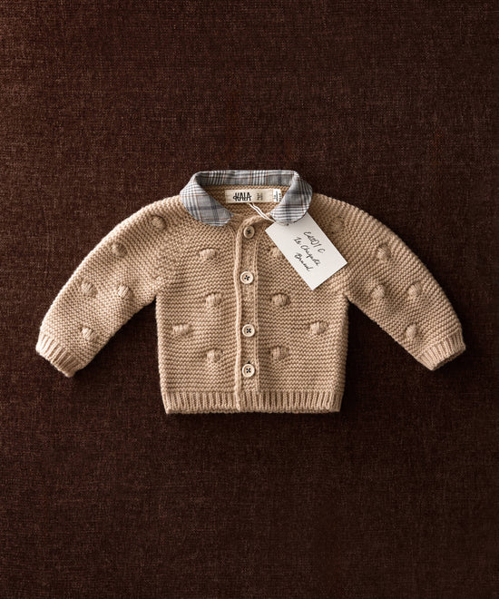 Chaqueta de punto de bebé beige con bodoques de punto y cuello Príncipe de Gales sobre fondo marrón