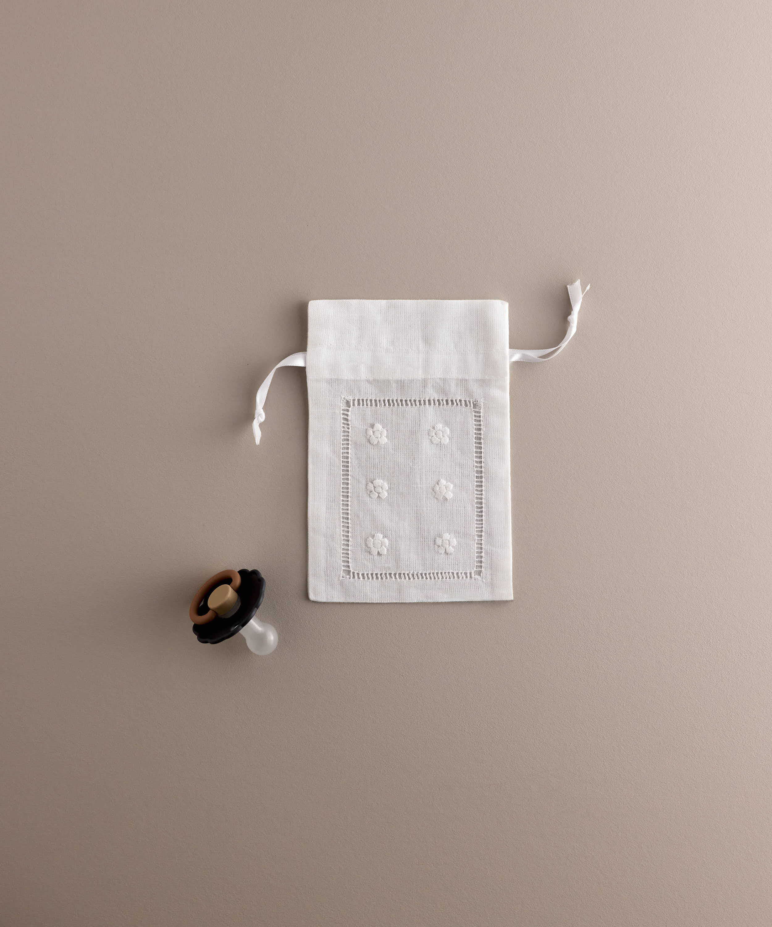 Porta chupete Tinylace en lino blanco bordado con flores y vainica, junto a chupete de referencia